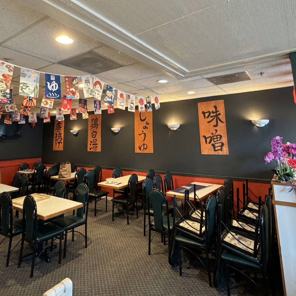 Japanese restaurant dining room with empty tables, hanging food banners, calligraphy wall scrolls, and floral arrangement.